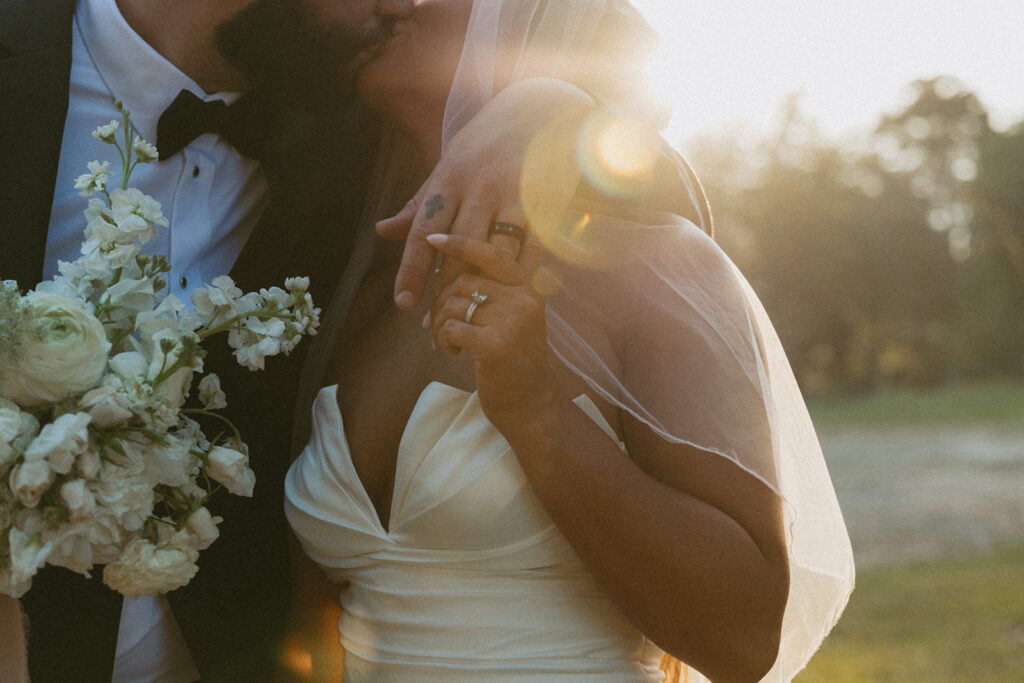 Man and woman in wedding attire kissing with the focus on their wedding rings and a sun flare. 