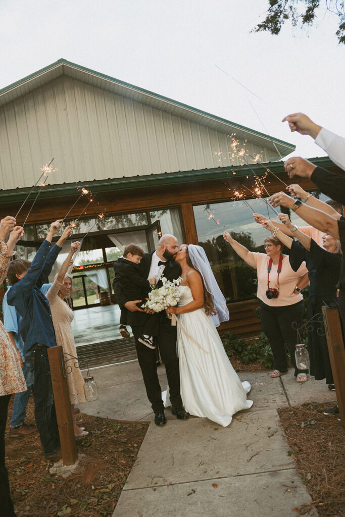 Man and woman in wedding attire walking through sparkler tunnel with their son.