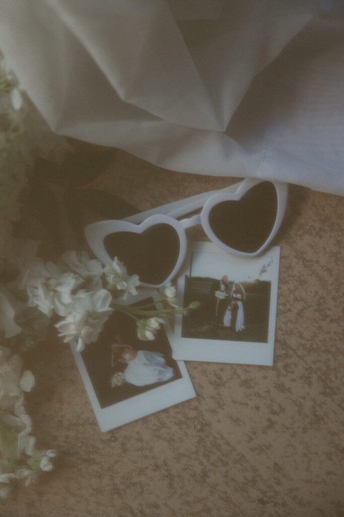 Flowers, wedding polaroids and white heart shaped sunglasses laying on the floor at wedding reception.