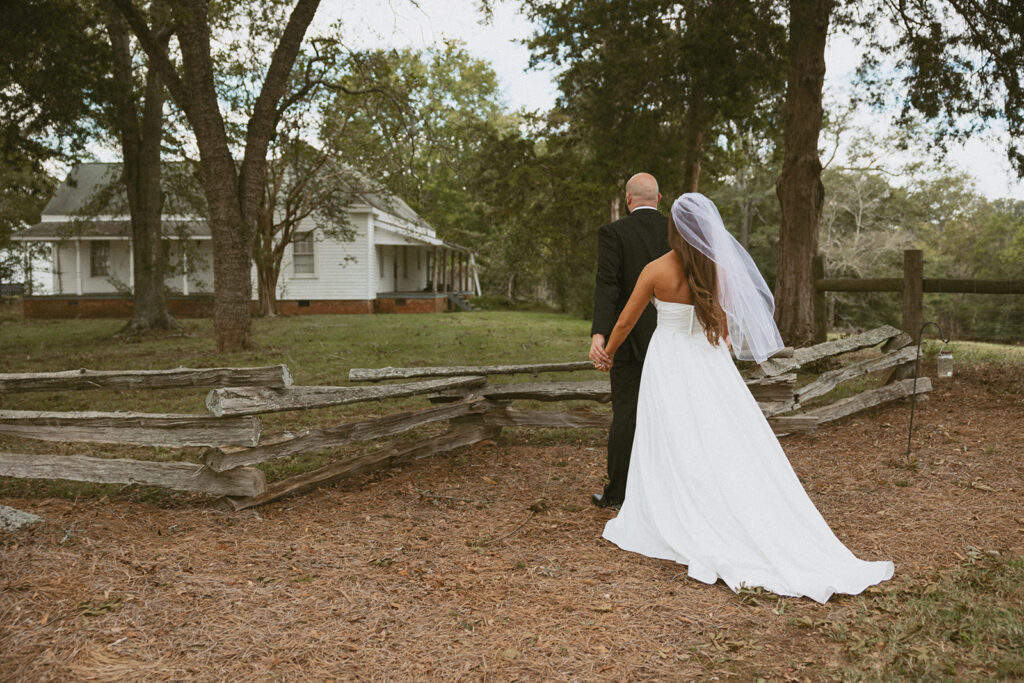 Woman in wedding dress standing behind man in a suit and holding his hands.