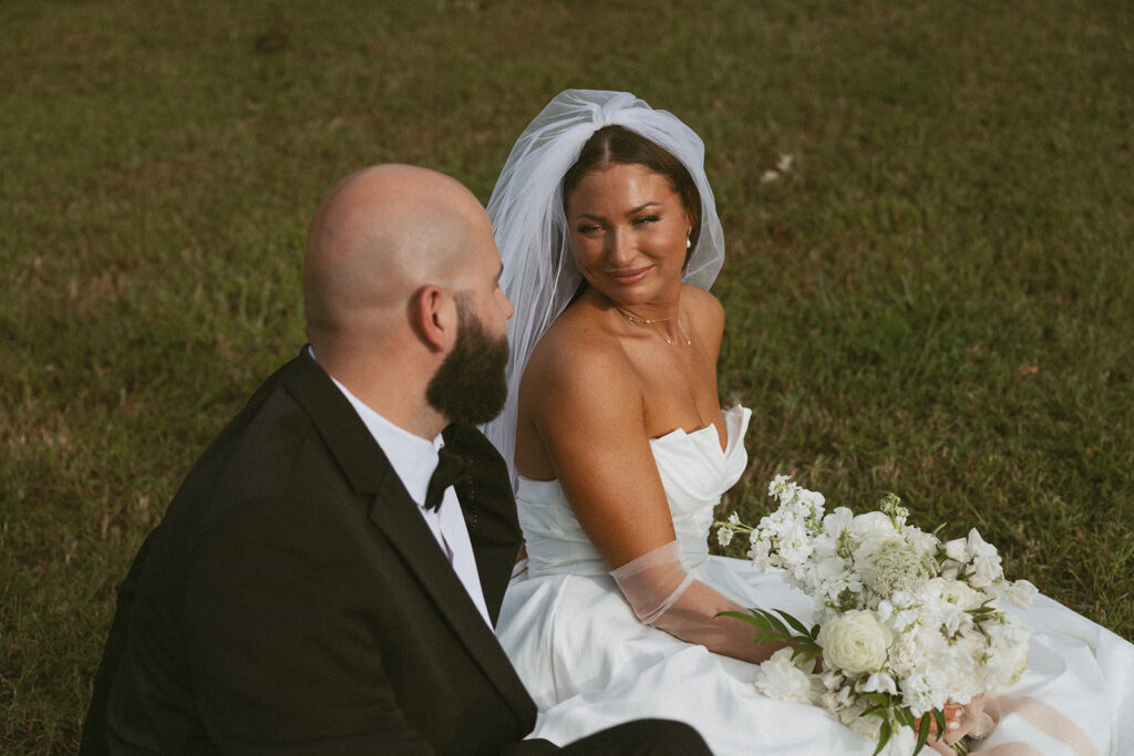 Man and woman in wedding attire sitting in grass and smiling at each other.