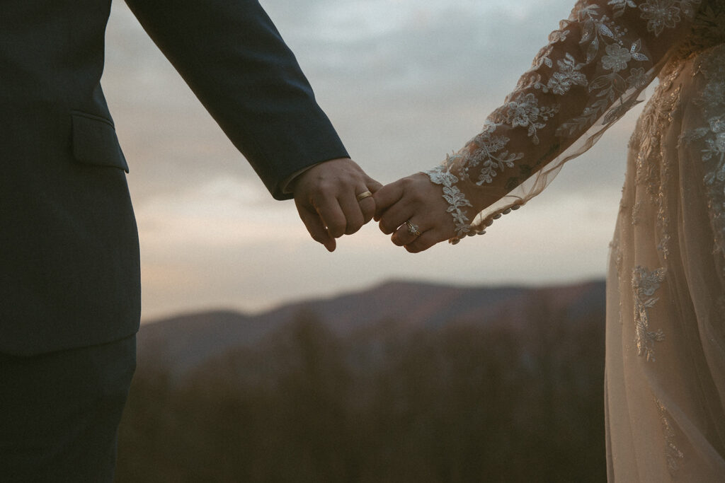 close up photo of bride and groom holding hands where you can see both of their wedding rings with mountains behind them