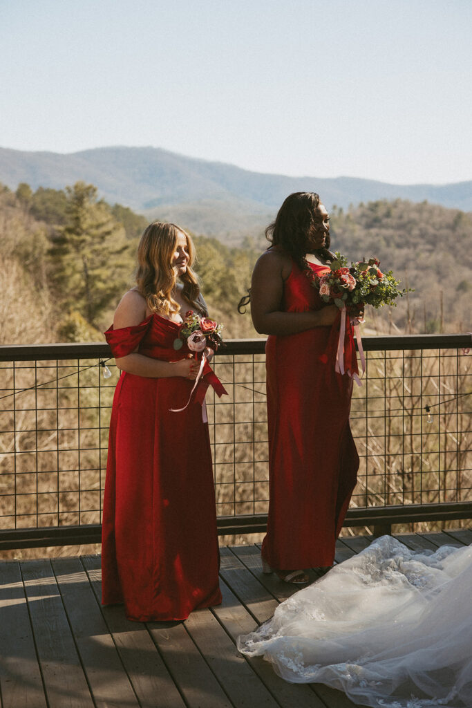 two bridesmaids in red dresses standing with bride during the ceremony on deck of airbnb with mountains behind them.