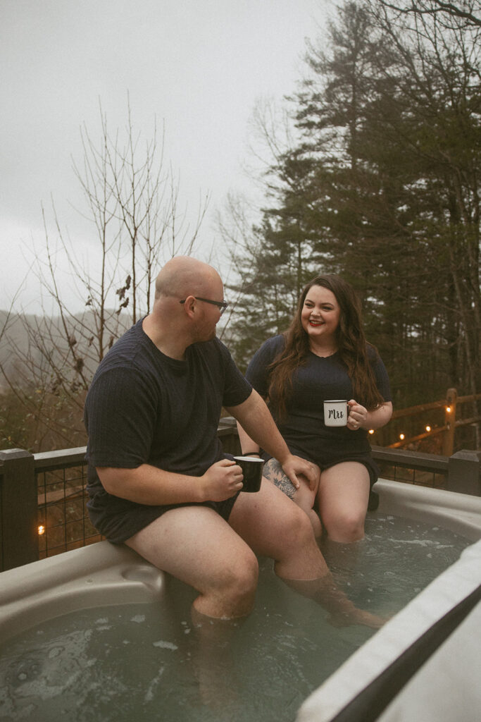 man and woman sitting on side of hot tub with their feet in, holding "his" and "hers" coffee mugs with mountains behind them.