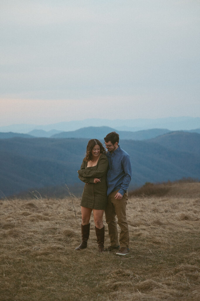 Man and woman cuddling and smiling at each other during engagement photos at Max Patch mountain in North Carolina.