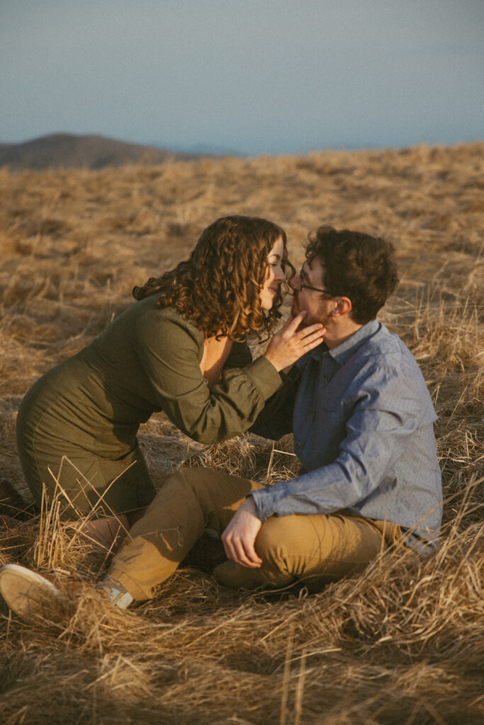Man and woman sitting on ground and going in for a kiss during engagement photos at Max Patch mountain in North Carolina.