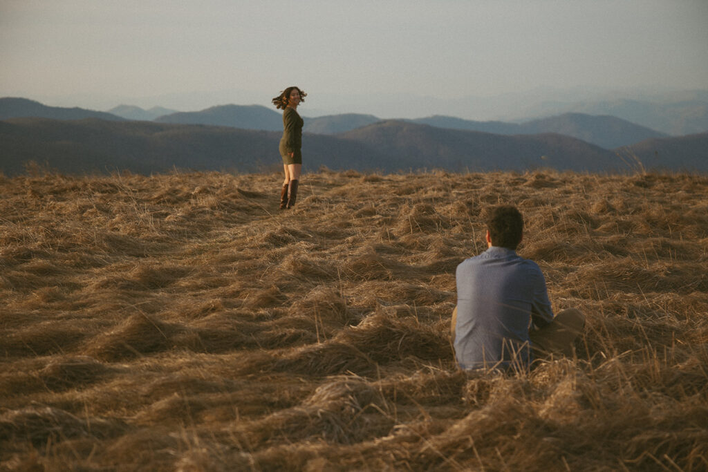 Woman walking away and looking back at man during engagement photos at Max Patch mountain in North Carolina.