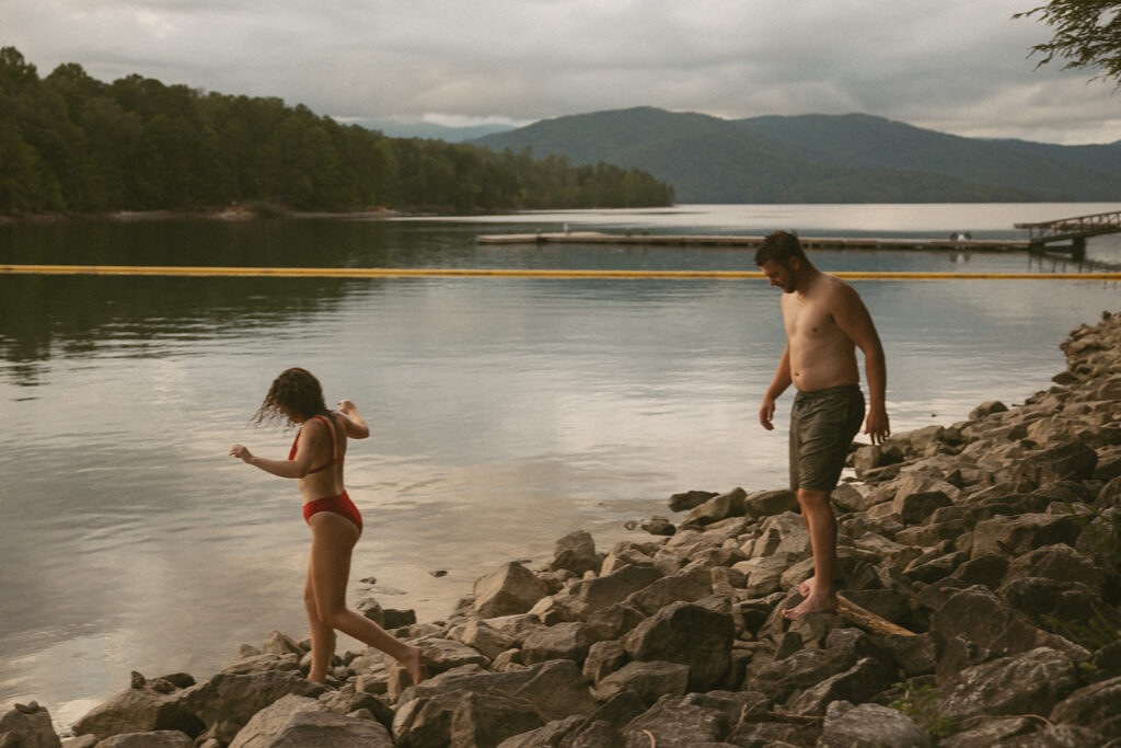 man and woman walking on rocks with lake and mountain behind them