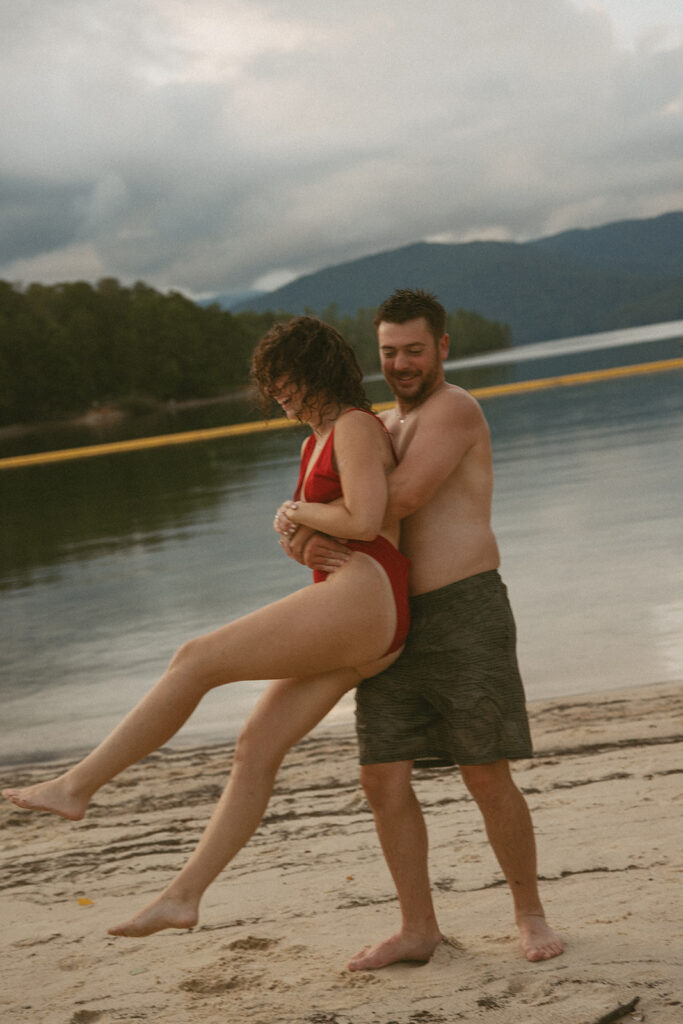 man spinning woman on a lake beach with mountains behind them
