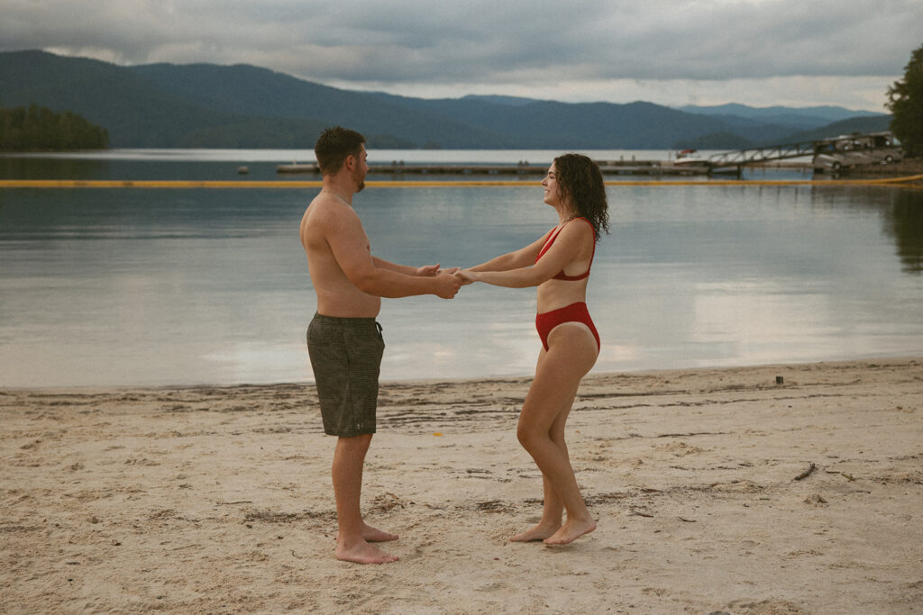 man and woman in swimsuits holding hands and spinning on a beach with the lake and mountains behind them
