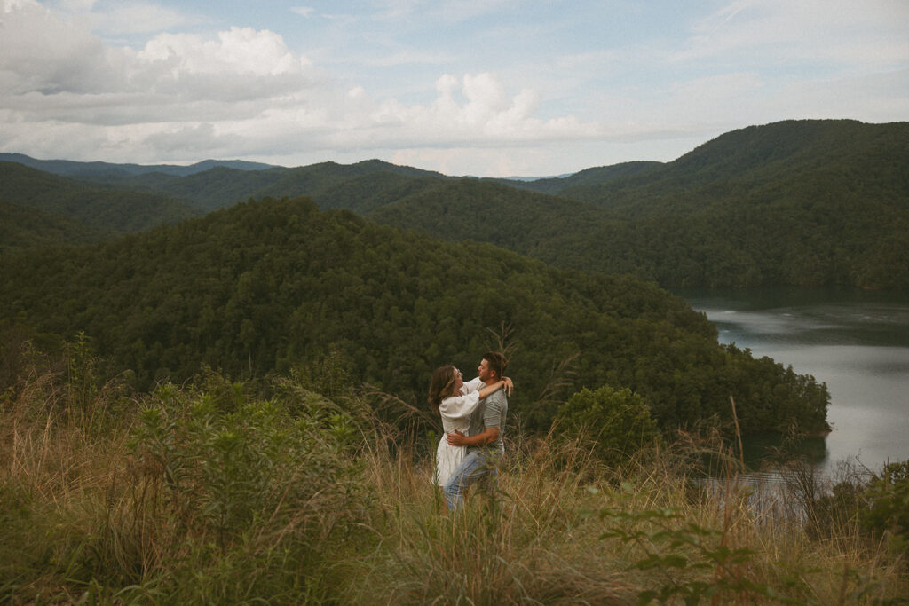 woman with arms wrapped over mans shoulders with mountains and lake behind them