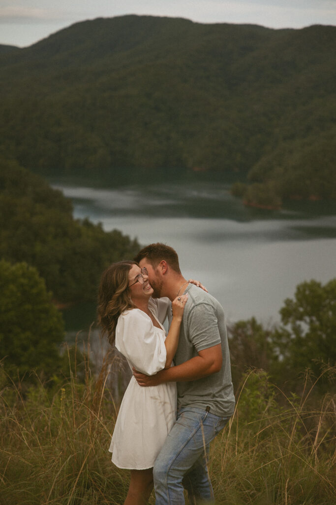 man and woman kissing and laughing during photoshoot with mountains behind them