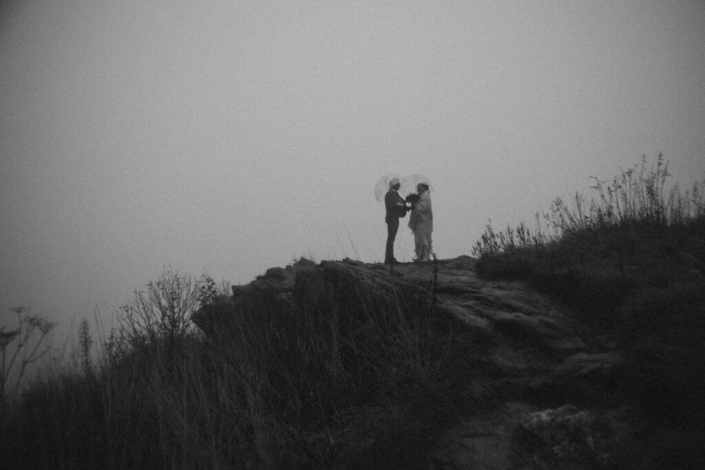Man and woman holding umbrellas standing a rock outcropping during their elopement