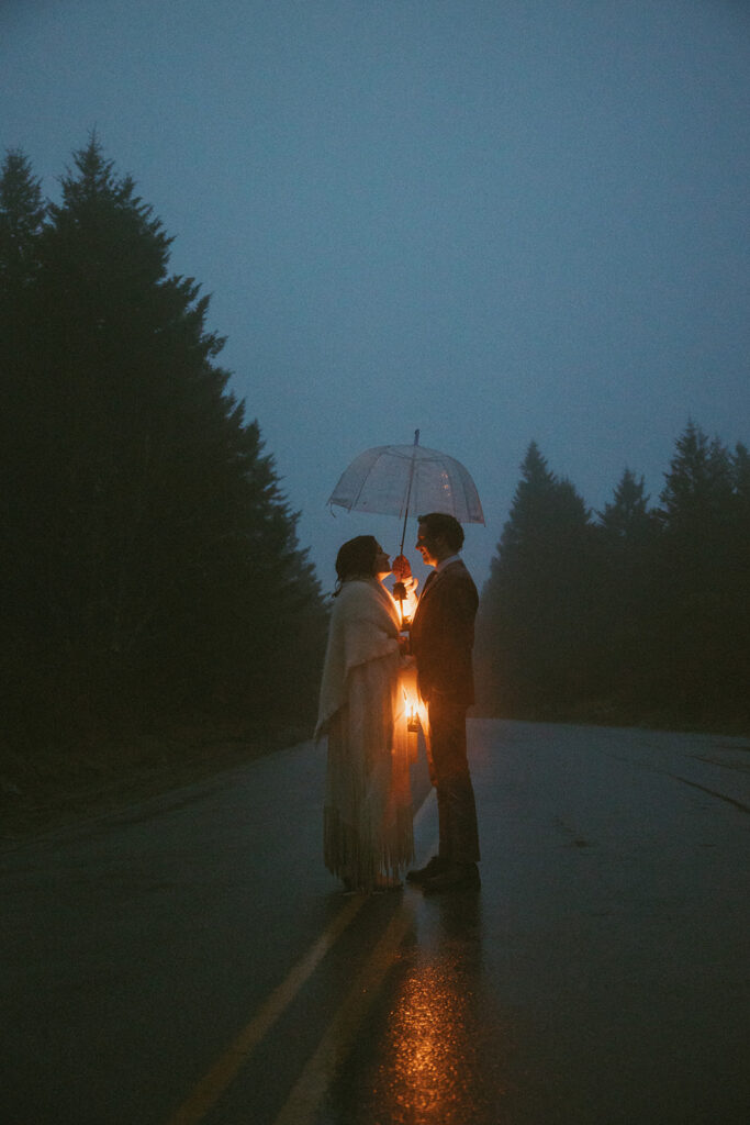 man and woman standing underneath umbrella on the road holding lanterns with trees behind them on their elopement day