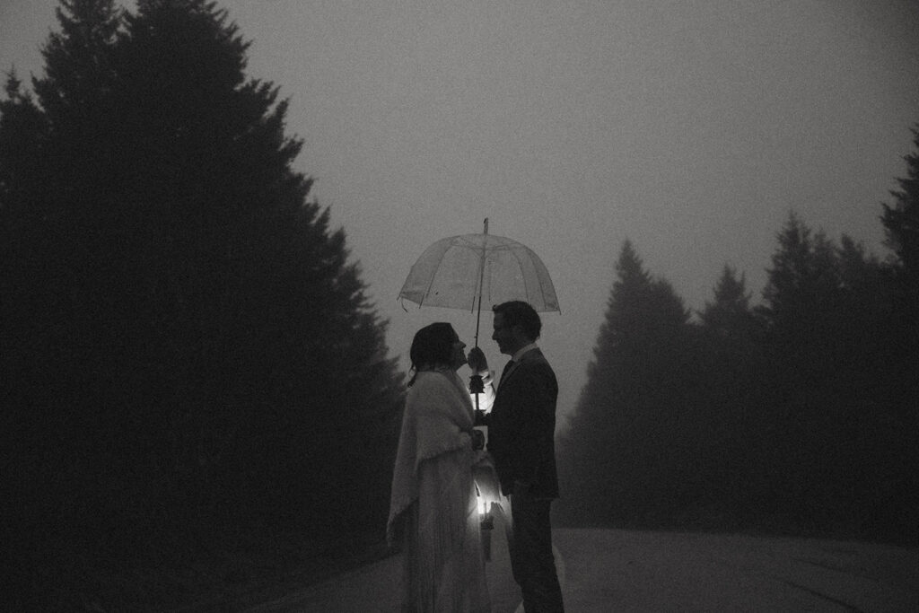 man and woman standing underneath umbrella on the road holding lanterns with trees behind them on their elopement day