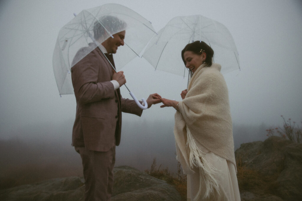 Man putting wedding ring on woman while they hold umbrellas during their rainy day mountain elopement