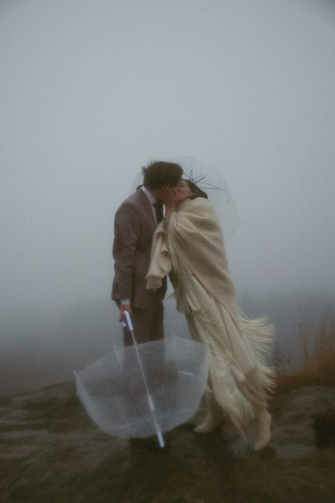 man and woman kissing while holding umbrellas during their mountain elopement
