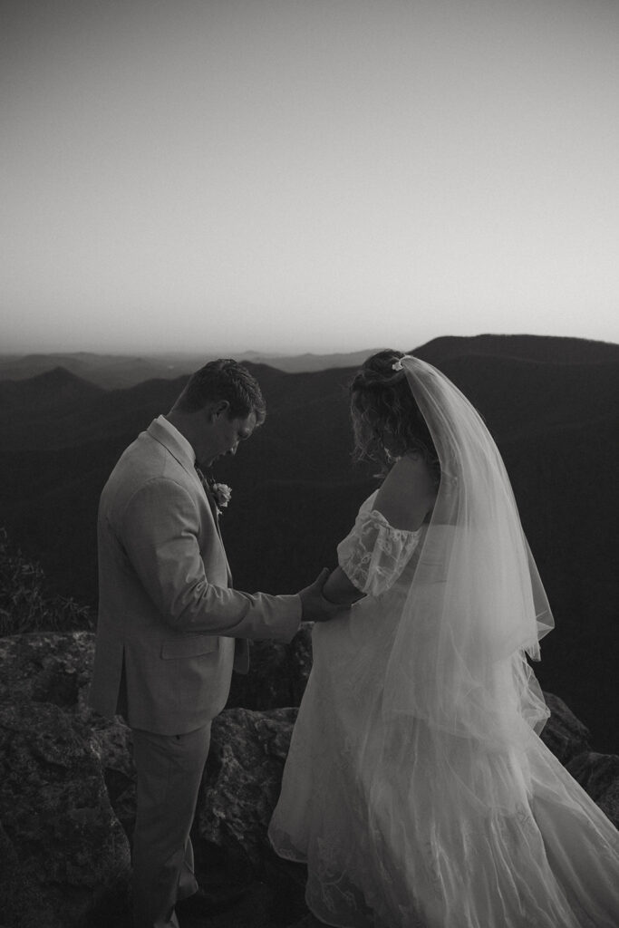 man help woman in wedding dress down a rock during hiking elopement in north carolina