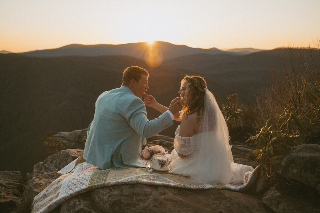 man and woman in wedding attire feeding each other cake with sun setting in front of them