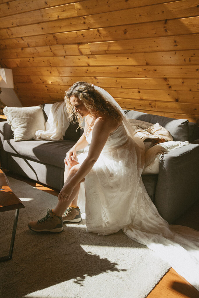 woman in wedding dress sitting on couch putting on her hiking boots for hiking elopement