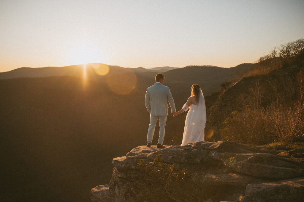 man and woman in wedding attire holding hands on a rock outcropping overlooking the north carolina mountains with the sun setting in front of them