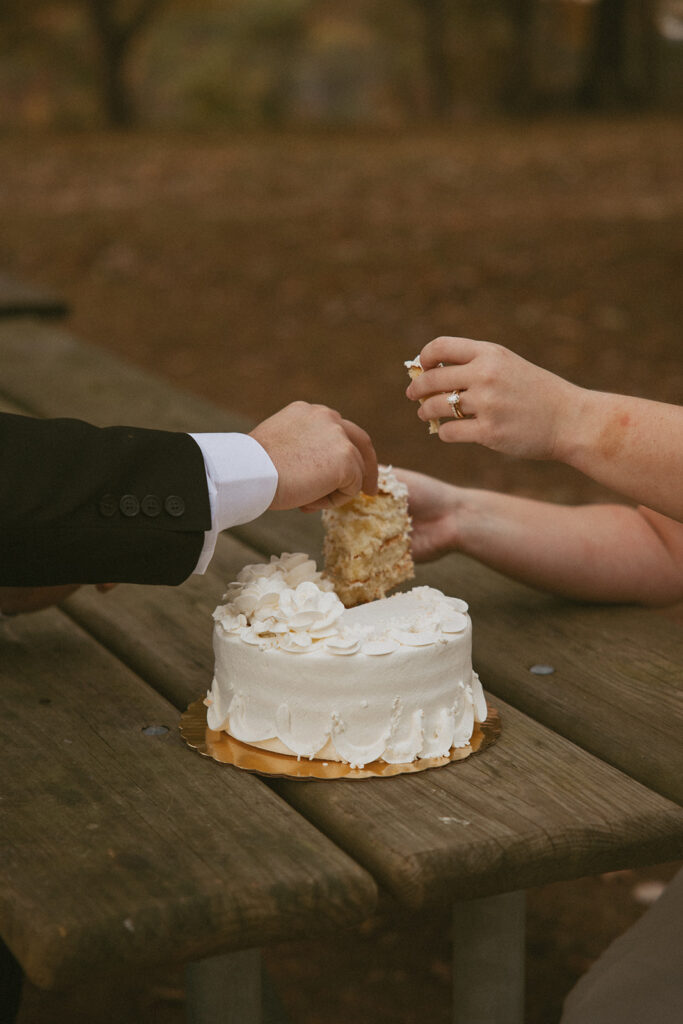man and woman sitting at picnic table feeding each other cake during their elopement